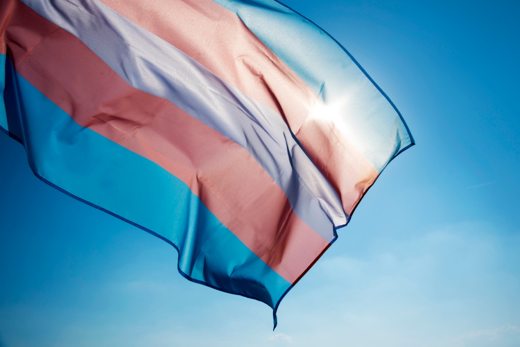 closeup of a transgender pride flag waving on the blue sky, moved by the wind, with the sun in the background